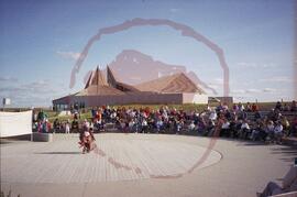 Stacy Spencer in traditional regalia performing in amphitheatre with audience and visitor centre ...