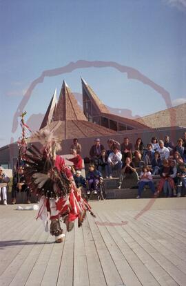 Stacy Spencer in traditional regalia performing in amphitheatre with audience and visitor centre ...